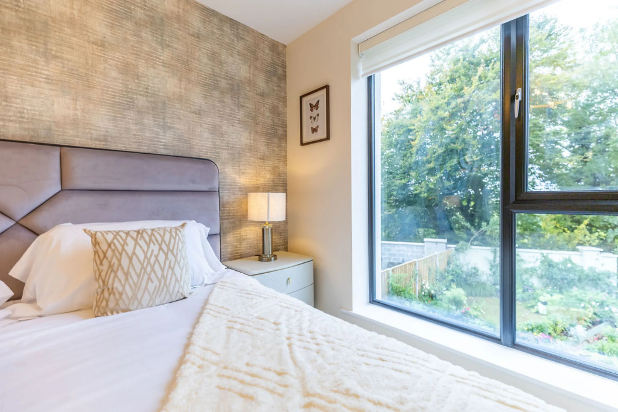 The image shows a bedroom interior with a bed, nightstand, and a large window overlooking a garden. The room has a neutral color palette with a textured wallpaper and a framed butterfly art piece.