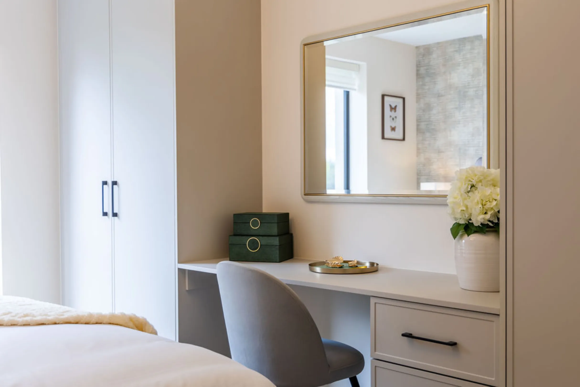 The image showcases a bedroom interior with a vanity area. The vanity features a mirror, desk, chair, and decorative items like boxes and flowers.