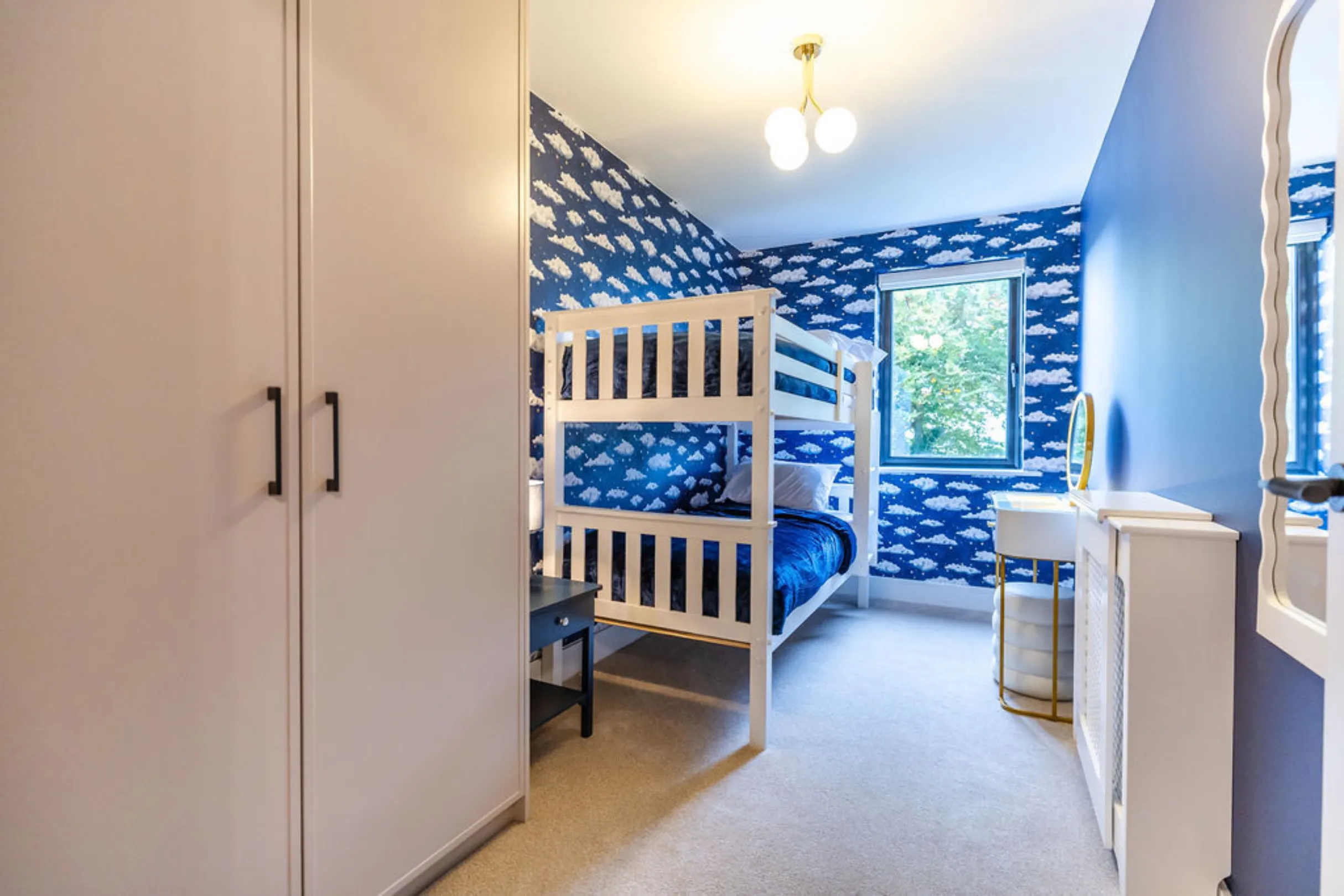 The image shows a child's bedroom with a white bunk bed, cloud-patterned wallpaper, and a small vanity. The room has a modern design with a blue and white color scheme.