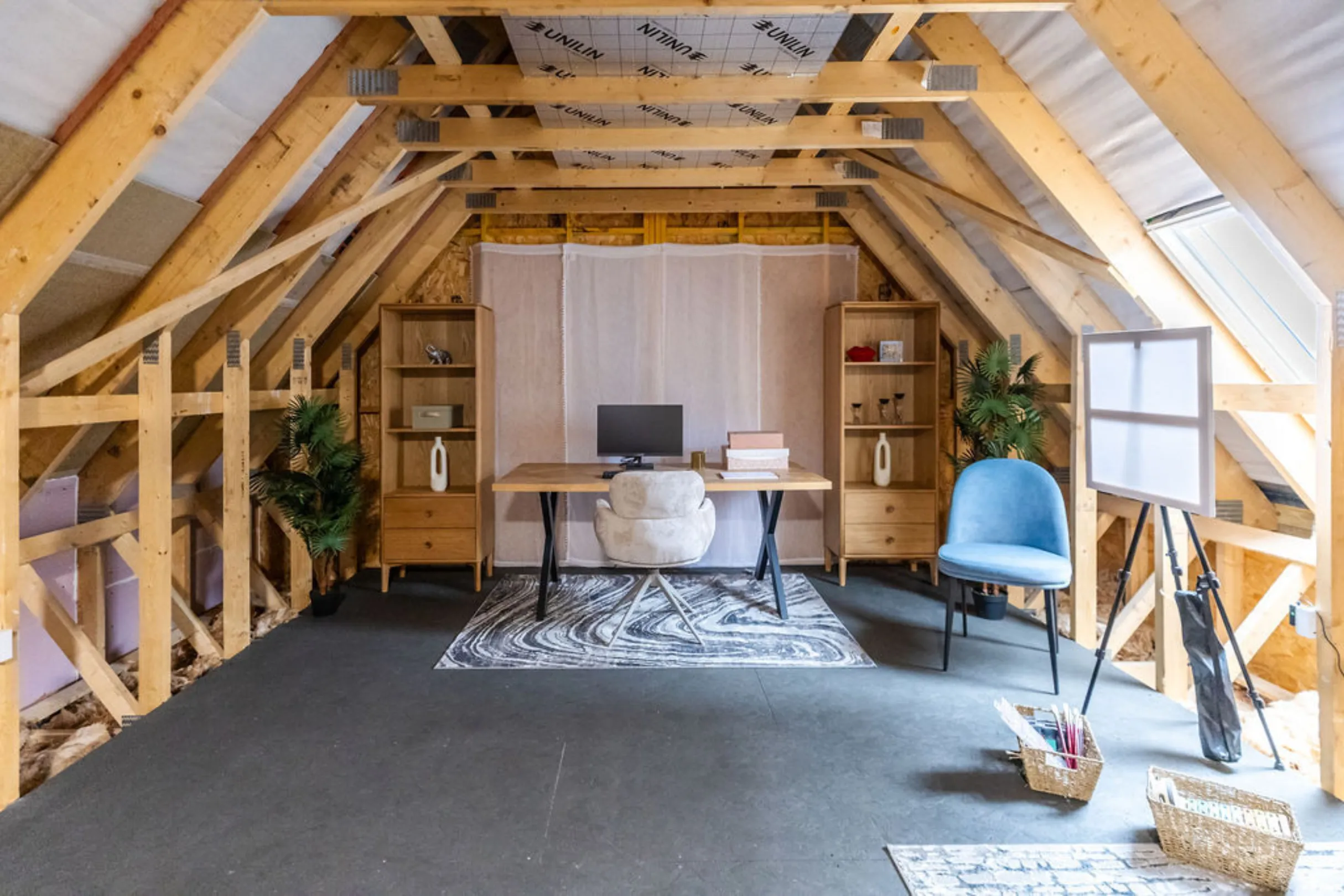 The image shows a newly renovated attic space designed as a home office. The room features a desk, bookshelves, and a sitting area with a modern aesthetic, set against the backdrop of exposed wooden beams.