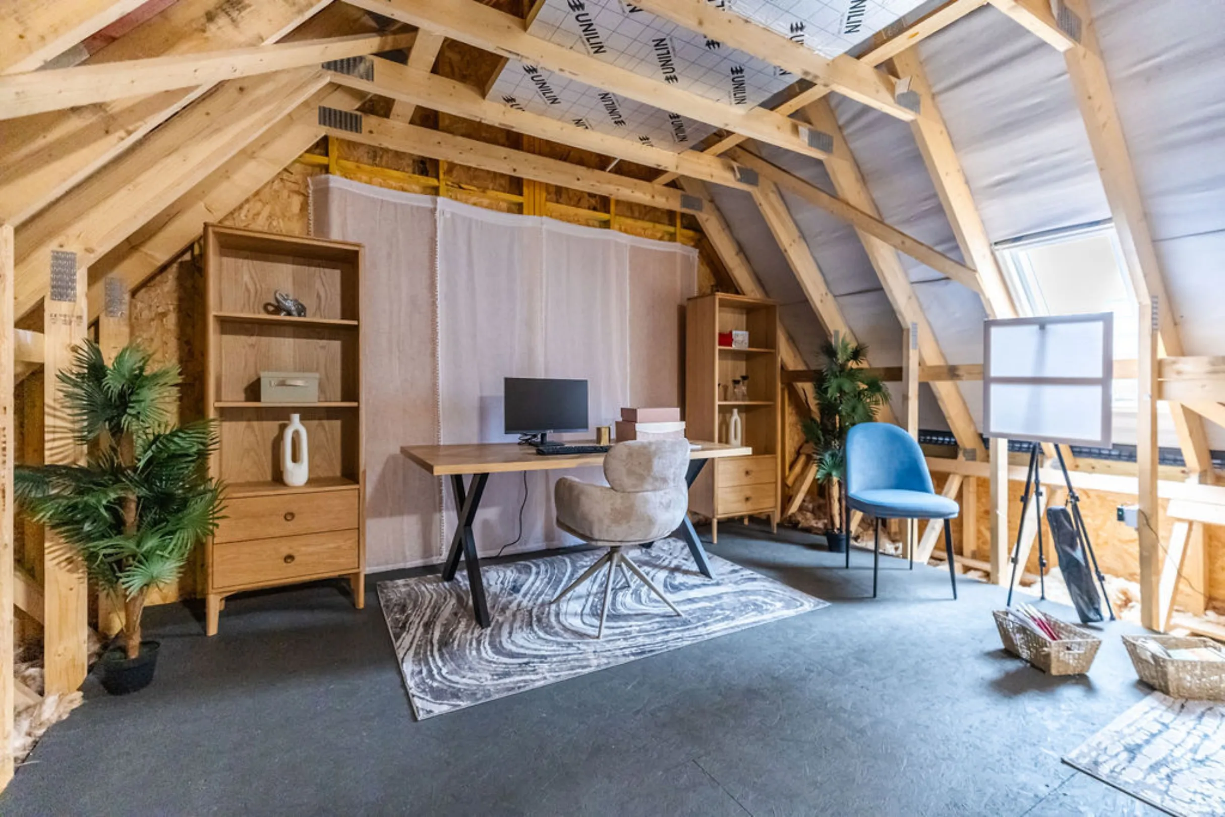 The image shows a home office in an attic space. The room features a desk with a computer, shelving units, a chair, and decorative plants.