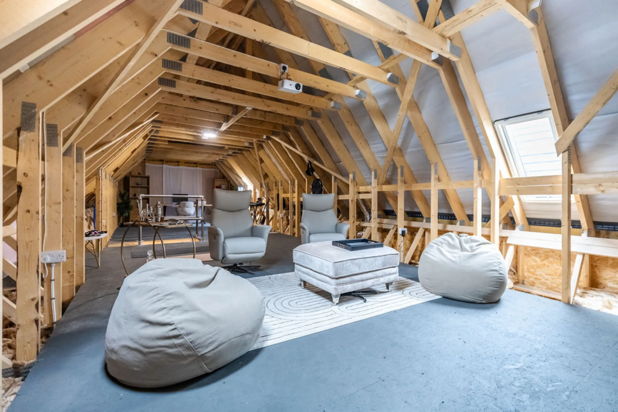 The image shows a renovated attic space converted into a modern living area. It features exposed wooden beams, comfortable seating, and a neutral color palette.