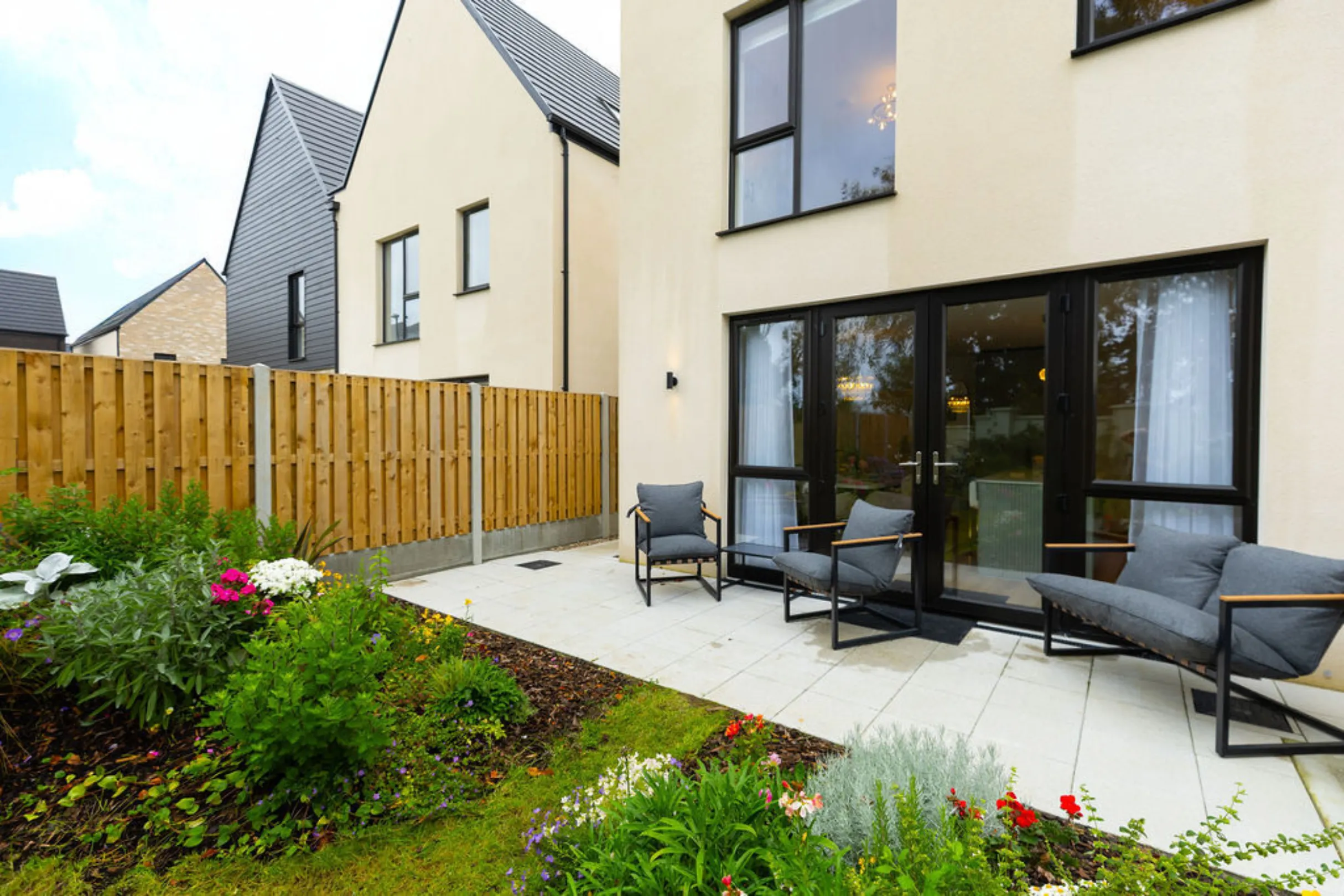 The image shows the exterior of a modern house with a patio and a garden. The patio has seating, and the garden is filled with plants and flowers.