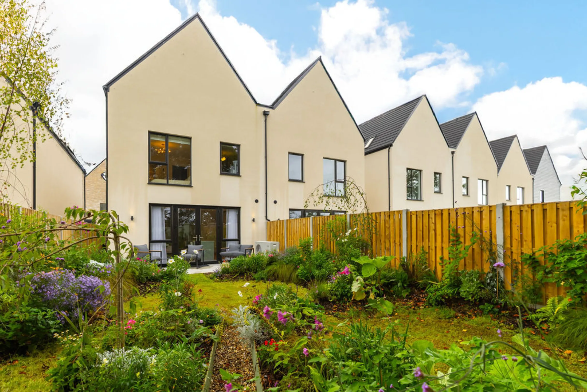 The image shows a row of modern houses with a well-maintained garden in the foreground. The houses have a simple, contemporary design with cream-colored walls and black roofs.