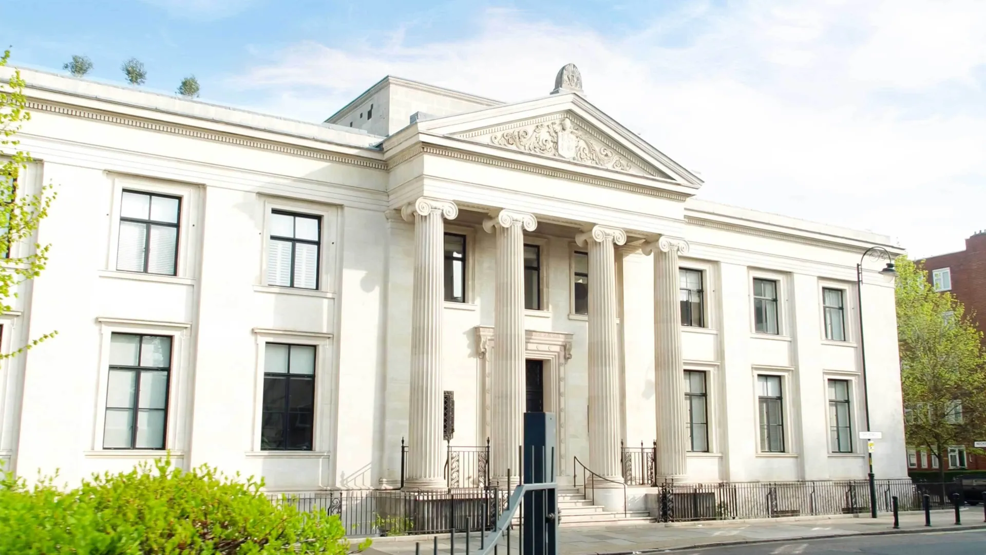 A imposing white building in the Italianate style with four columns supporting a triangular pediment with tympanum above the entrance. trees and bushes can be seem at the side.