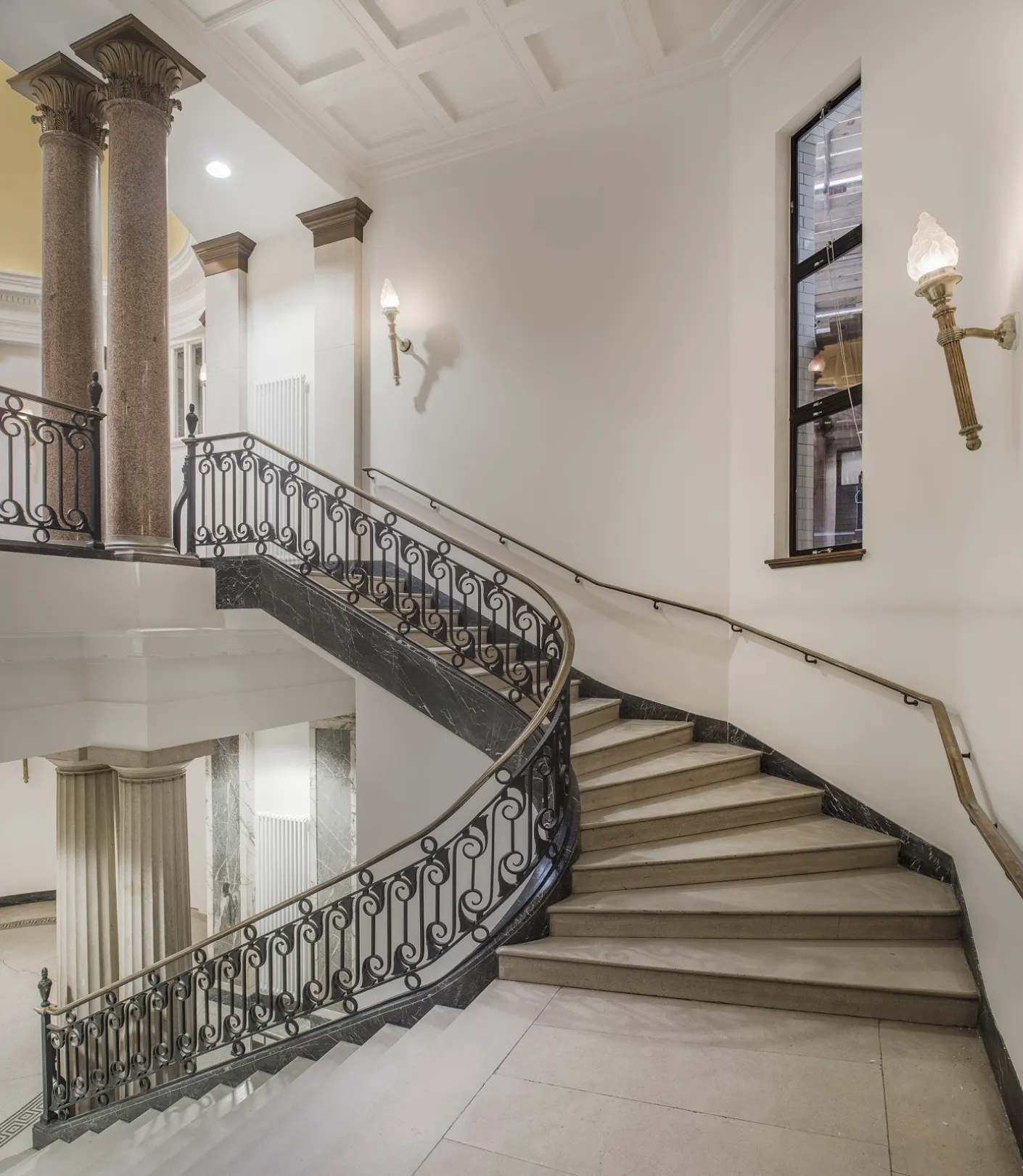 Grand curving staircase leading up to a second floor with marble columns and flame-style wall lights and an ornate balustrade