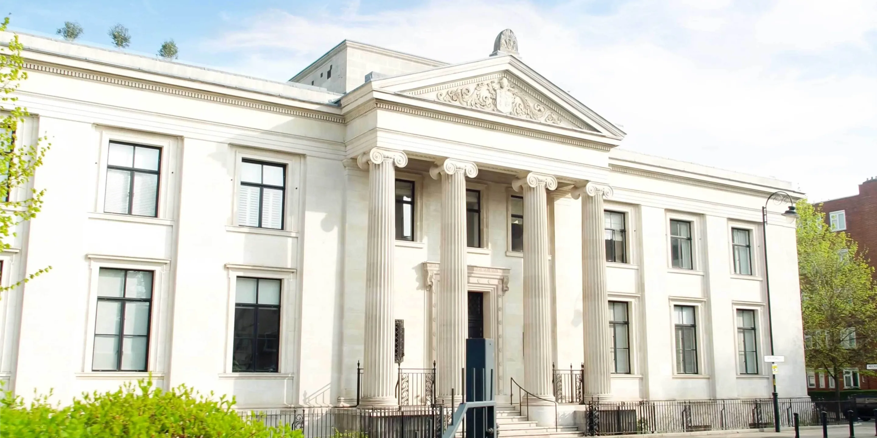 A imposing white building in the Italianate style with four columns supporting a triangular pediment with tympanum above the entrance. trees and bushes can be seem at the side.