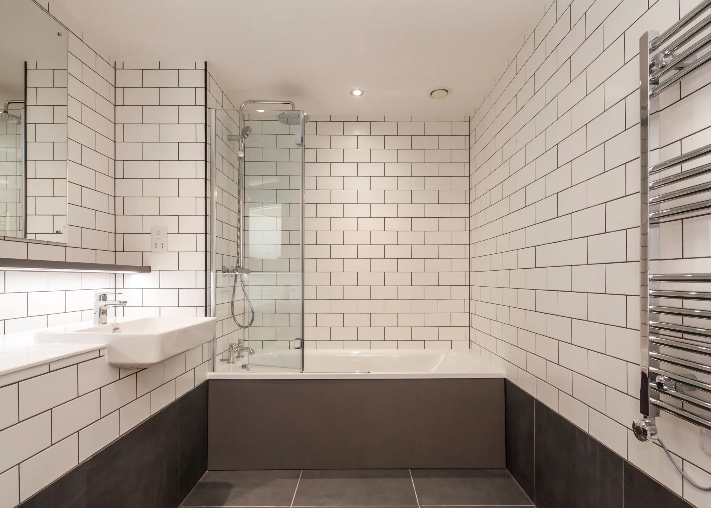 A bathroom with large format grey wall and and floor tiles.  White brick style wall tiles above the grey tiles, a semi-recessed white basin with chrome tap.  A bath with shower over head and a large wall mirror.