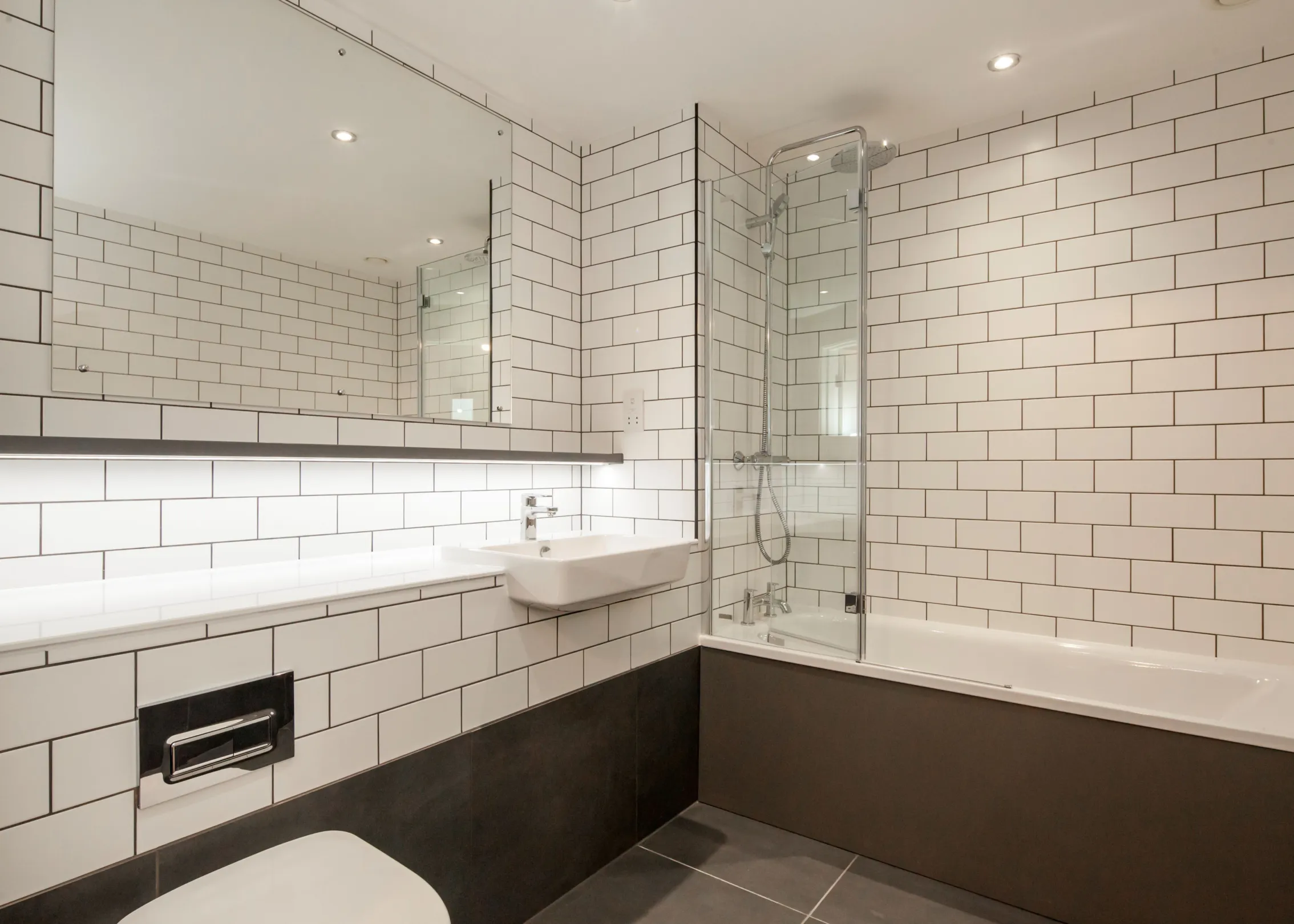 A bathroom with large format grey wall and and floor tiles.  White brick style wall tiles above the grey tiles, a semi-recessed white basin with chrome tap.  A bath with shower over head and a large wall mirror.