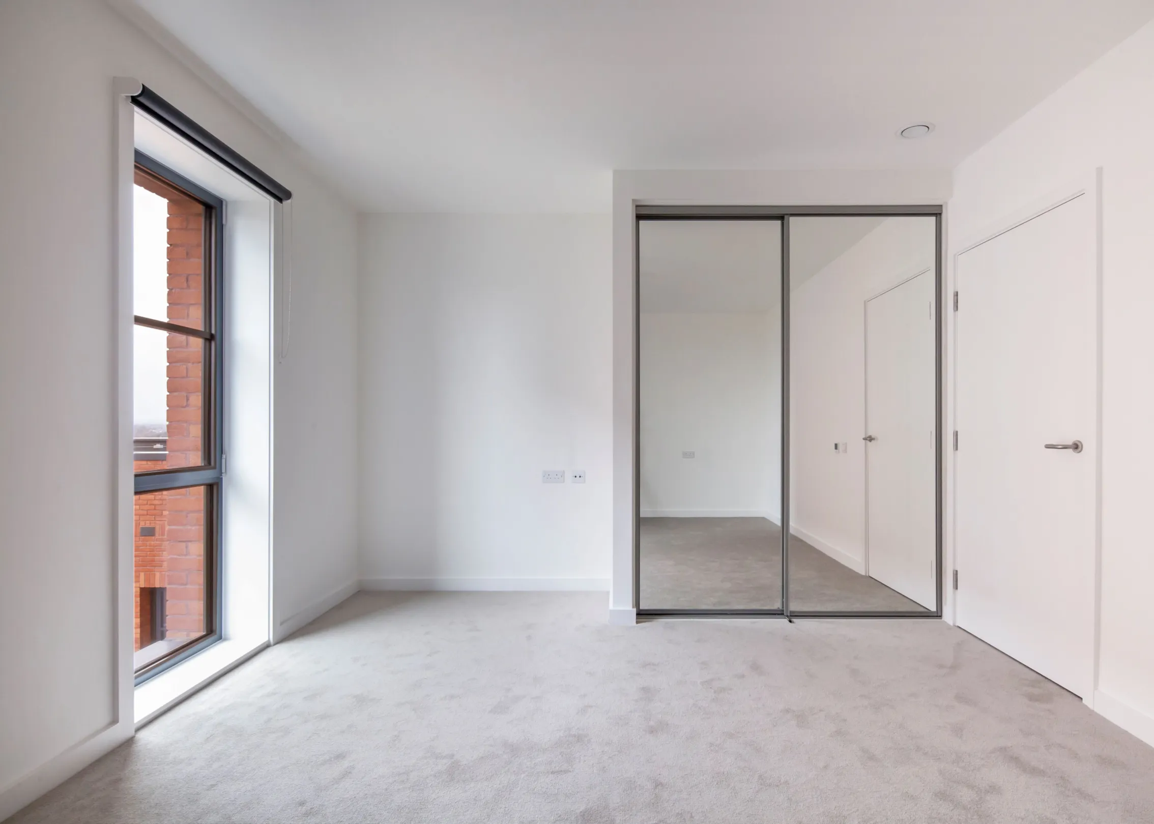 A bedroom in a new development, grey coloured carpet, sliding door wardrobe with mirrors and floor to ceiling height windows.
