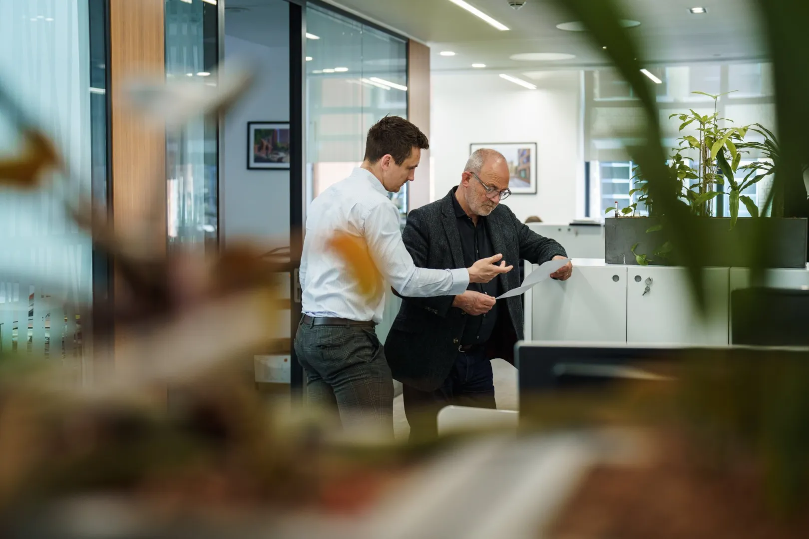 Two men discuss a document in a shared space of a modern office, with lots of internal planting and large glass partitions