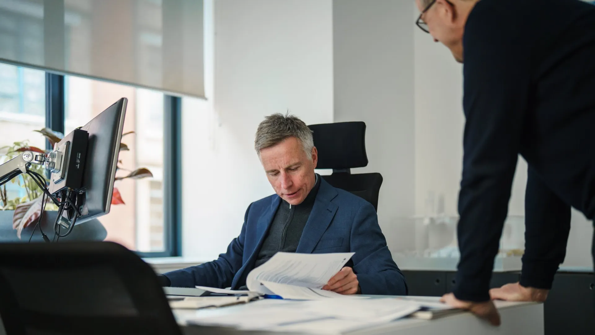 A man in a suit sits at a desk, with another man leaning in to discuss a document. A bright modern window is in the background.