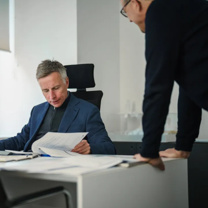 A man in a suit sits at a desk, with another man leaning in to discuss a document. A bright modern window is in the background.
