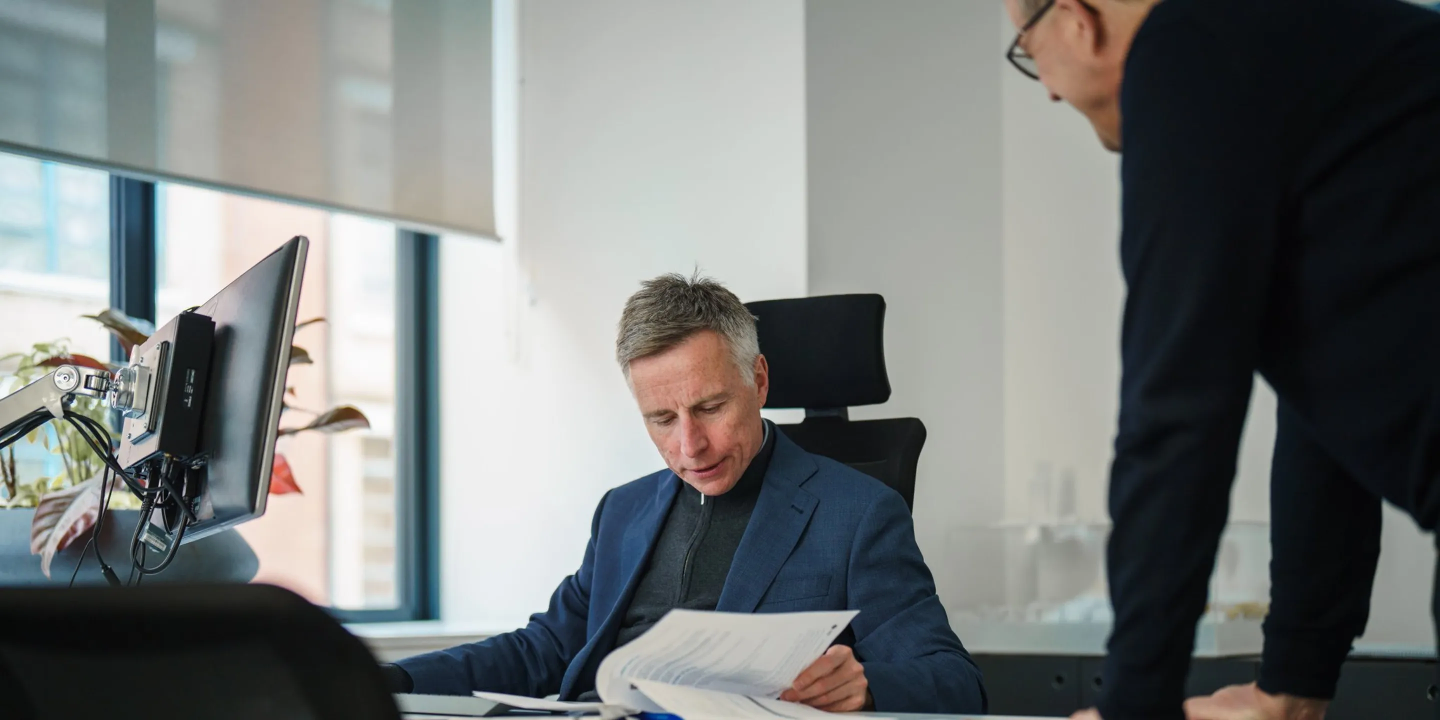 A man in a suit sits at a desk, with another man leaning in to discuss a document. A bright modern window is in the background.
