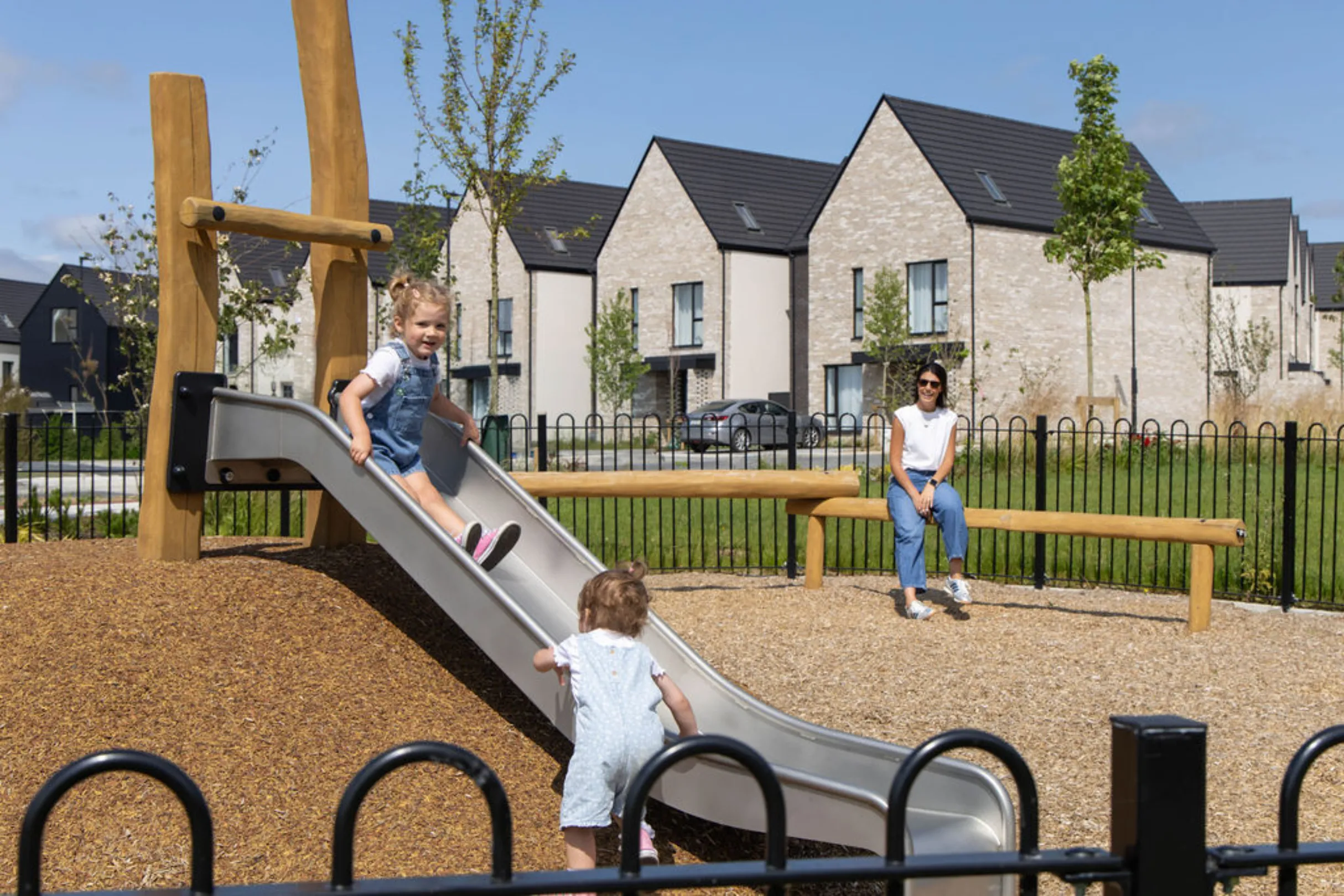 Two young children playing on a slide in a playground, with a woman sitting on a wooden bench and rows of houses behind.