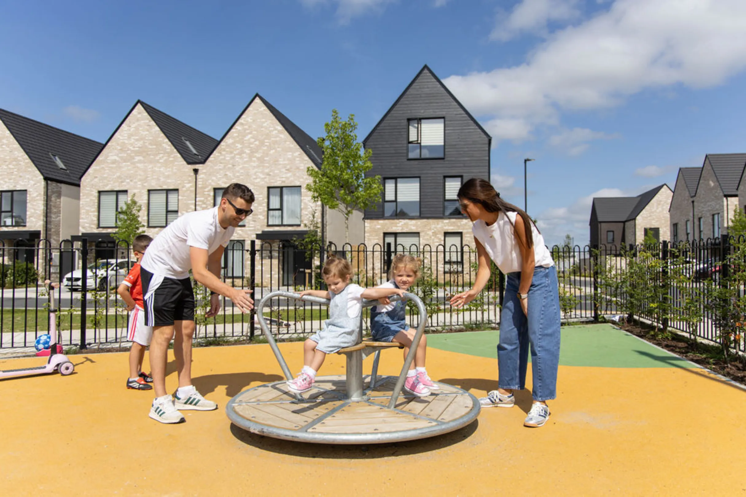 A family with children playing on a roundabout in a playground, with houses and a black-clad gable-fronted home in the background.