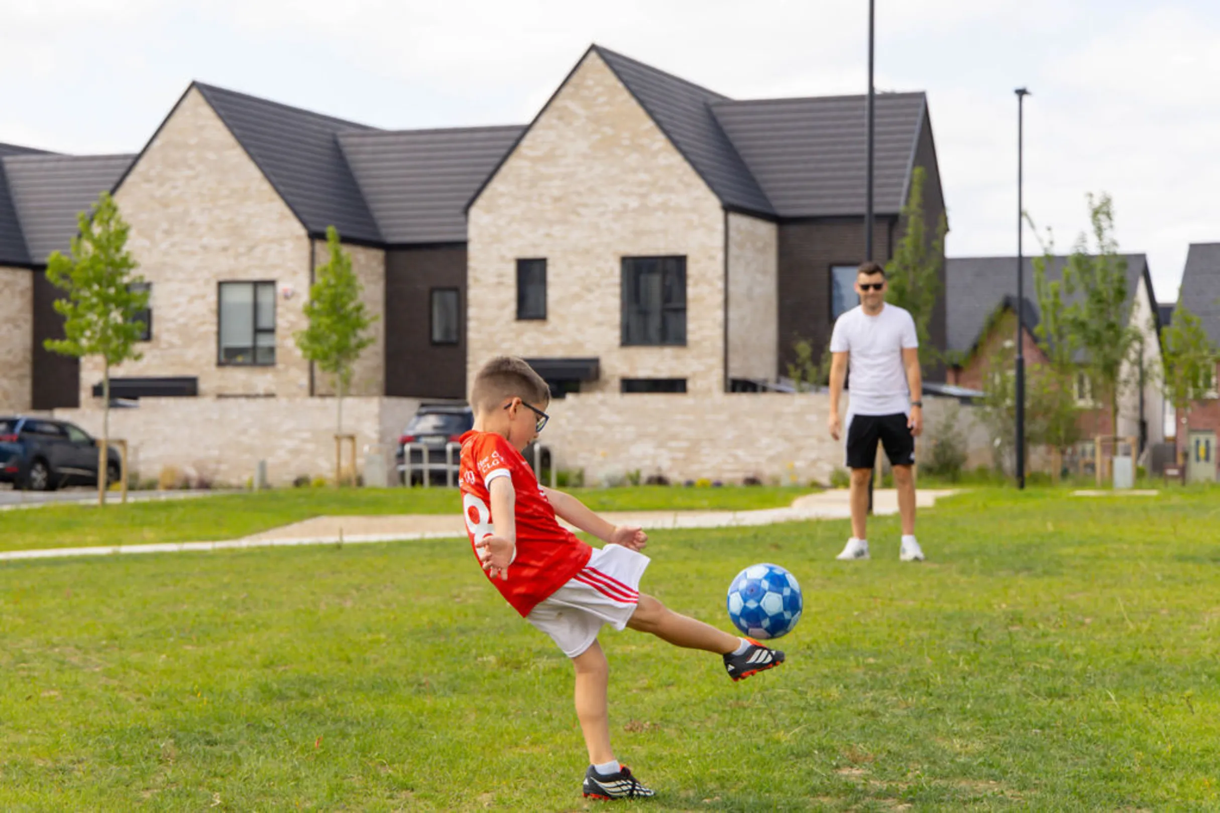 A boy in a GAA jersey kicking a football on open green space while a man looks on, with houses and parked cars behind.