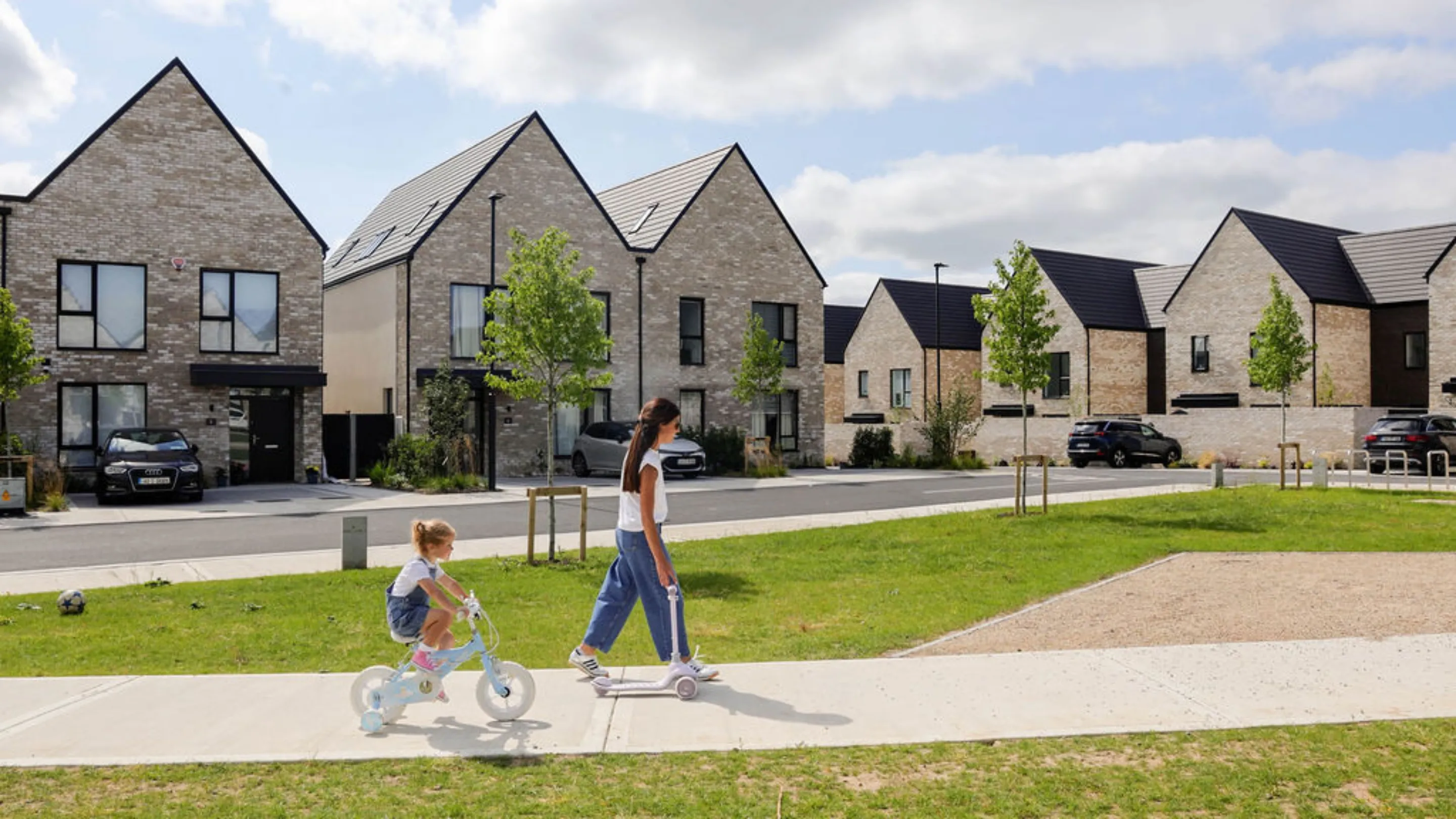 A woman walking with a young girl on a scooter and a young girl on a bicycle along a footpath, with a row of houses and parked cars alongside.