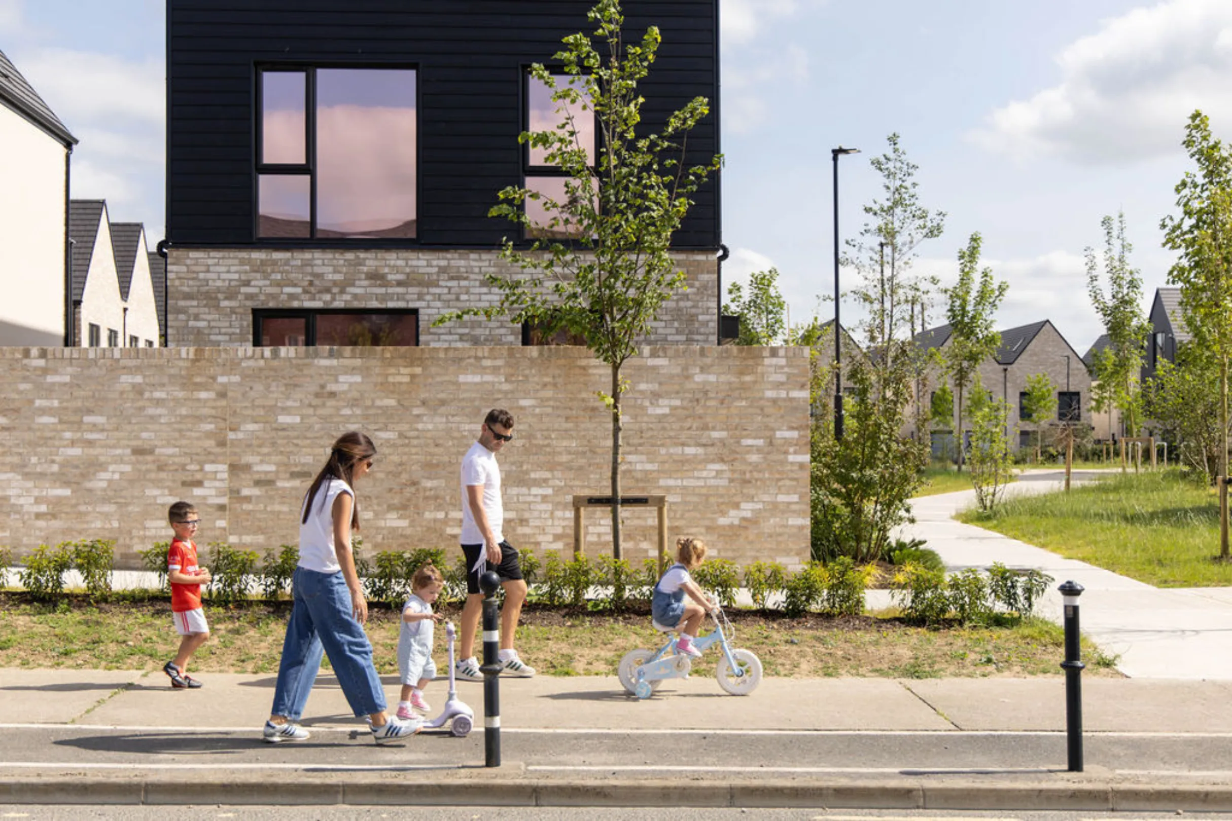 A family of five walking along a footpath past a boundary wall, with one child on a scooter and another on a bicycle, and a black-clad house behind.