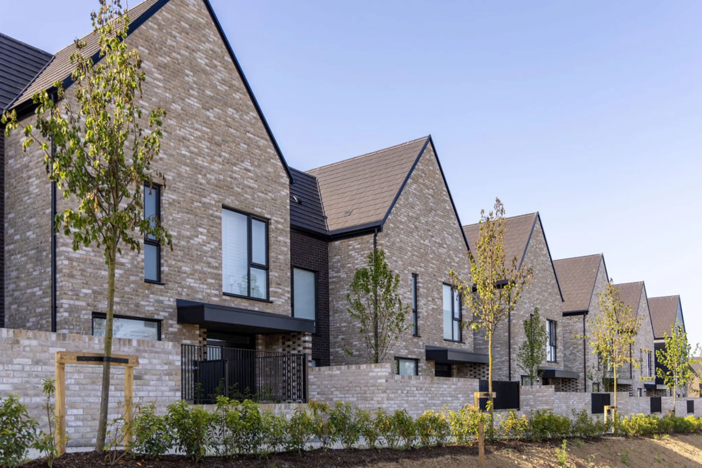 Close-up view of light brick, gable-fronted houses with dark roofs and black-framed windows, with young trees planted along the boundary wall.