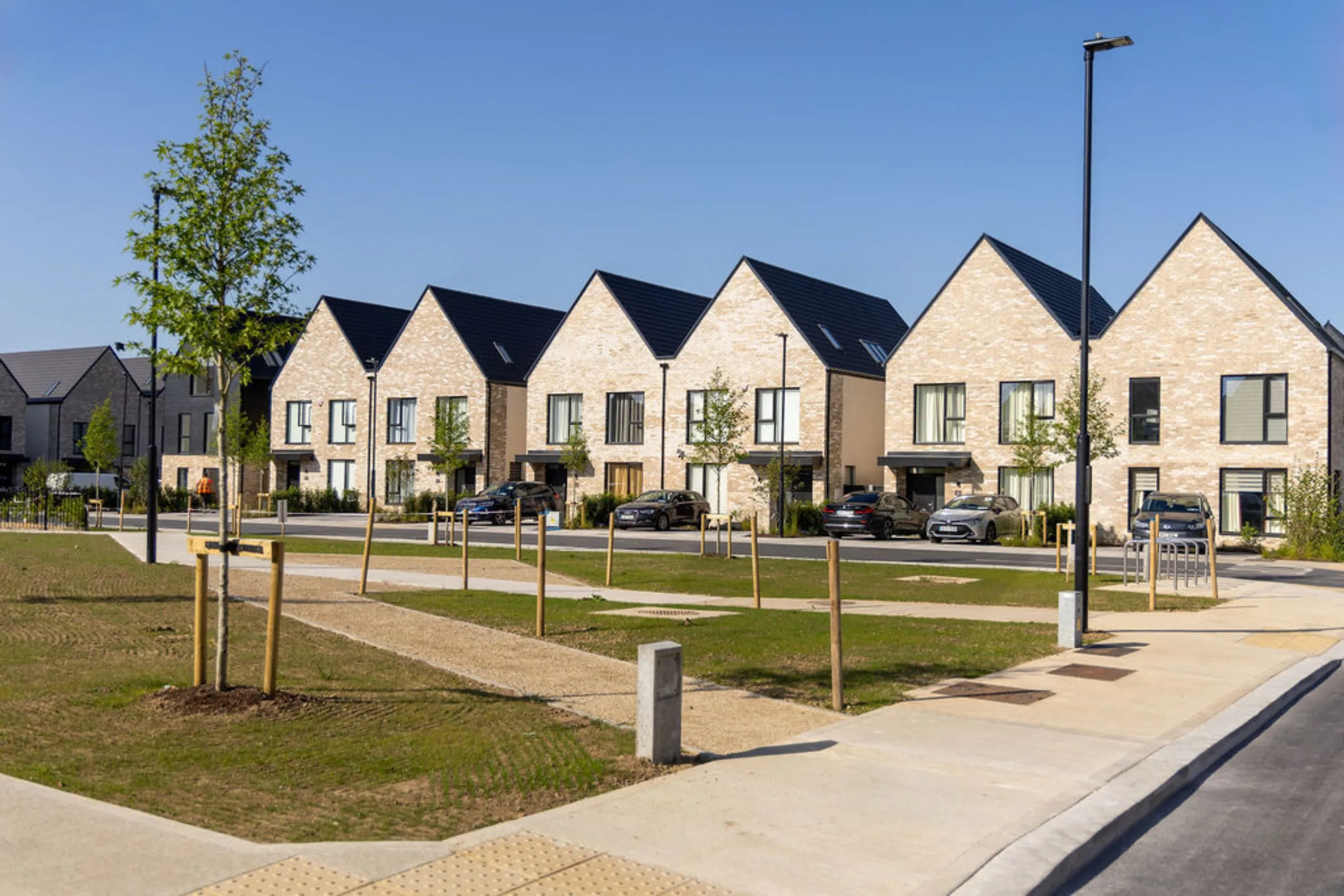 Row of light brick, gable-fronted houses facing a landscaped green space with footpaths, young trees and cars parked on the street.