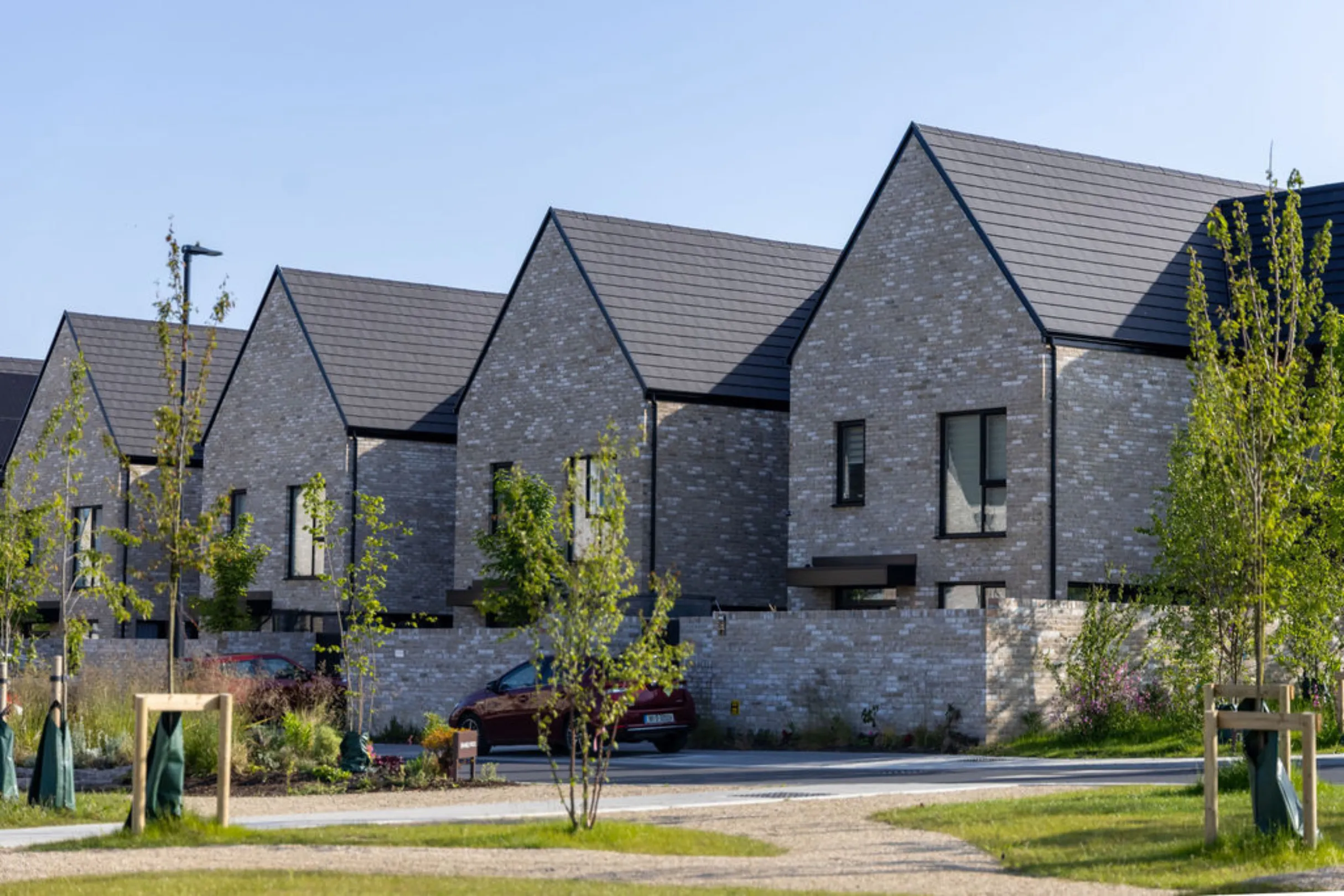 Row of grey brick, gable-fronted houses with dark roofs, seen from a green space with young trees and a car parked on the street.