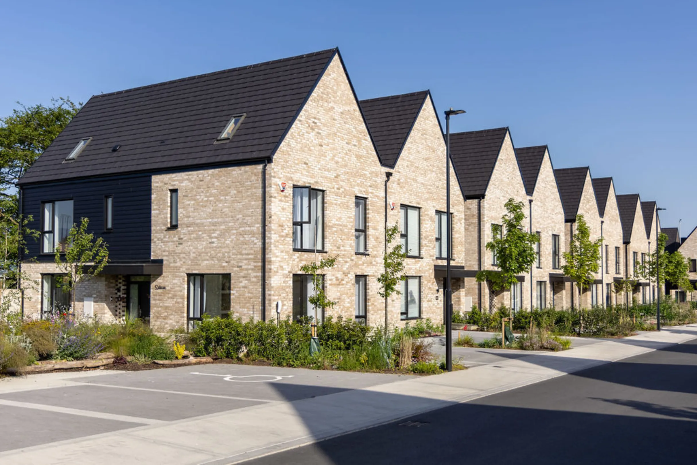 Row of two-storey brick houses with dark gabled roofs and a black weatherboarded end house, with front gardens and a disabled parking bay on the street.